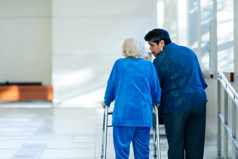 Pflegekraft begleitet eine Seniorin mit Gehhilfe durch einen hellen Flur